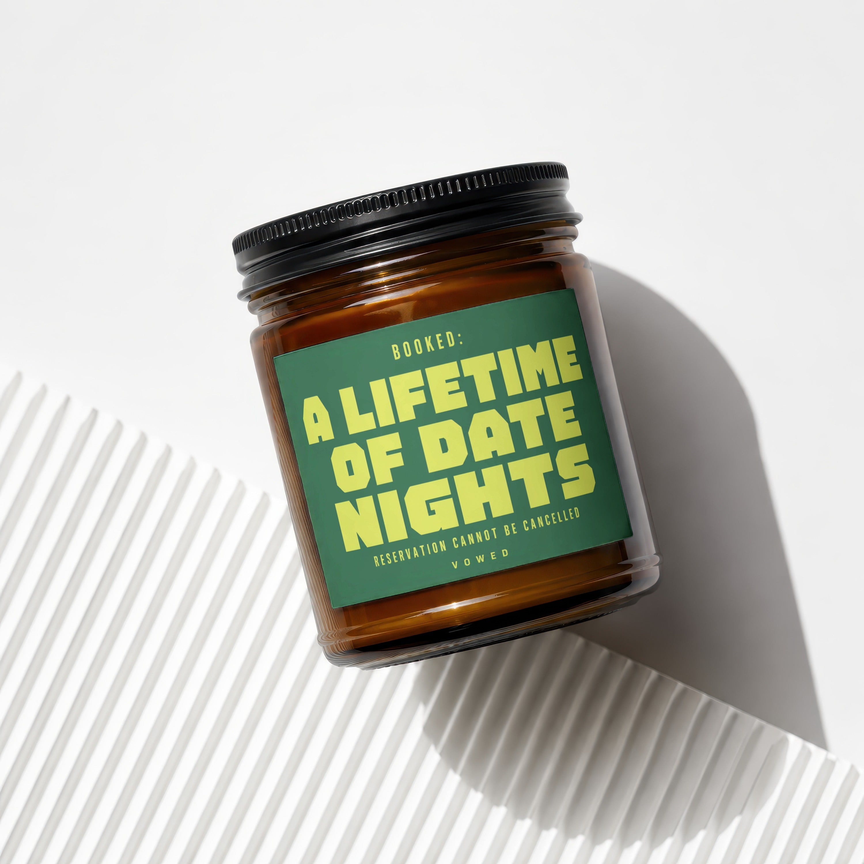 Amber candle with black lid and green label reading "BOOKED: A LIFETIME OF DATE NIGHTS".