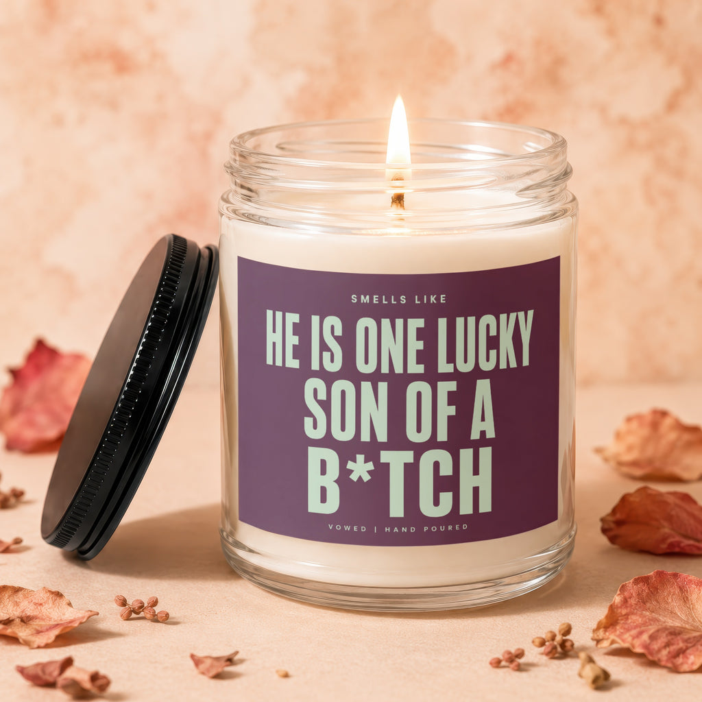 Lit candle in a glass jar with a purple label reading "HE IS ONE LUCKY SON OF A B*TCH".