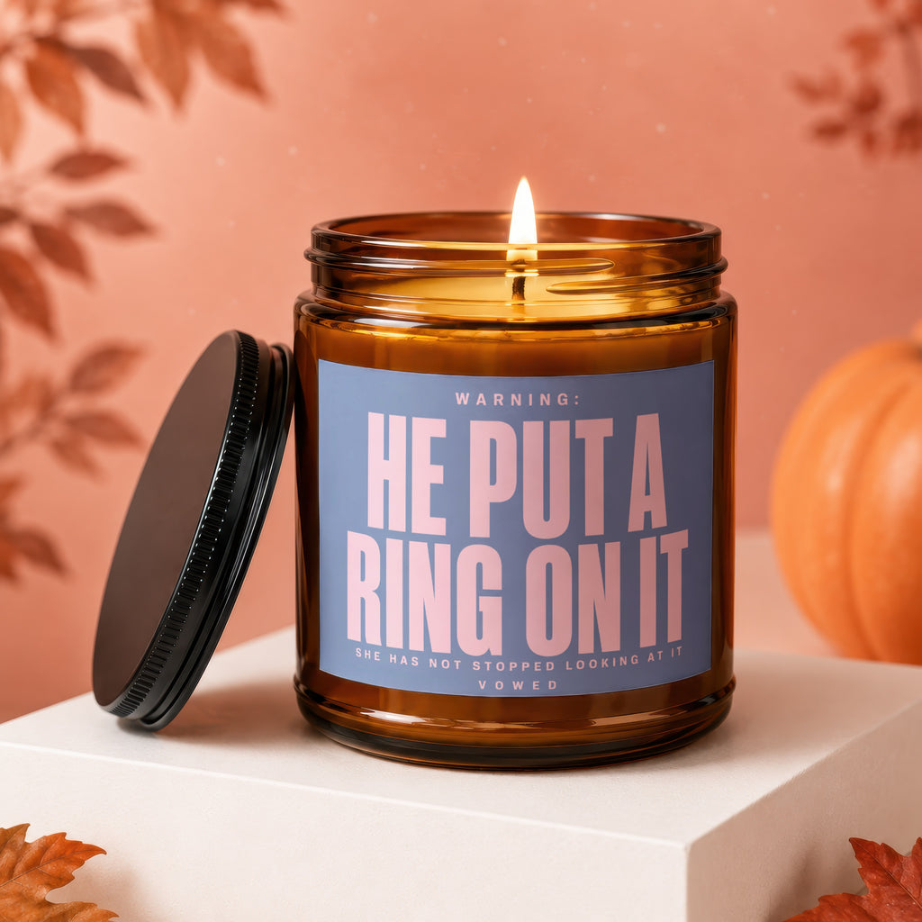 Amber jar candle with lit flame, purple label reading "HE PUT A RING ON IT."