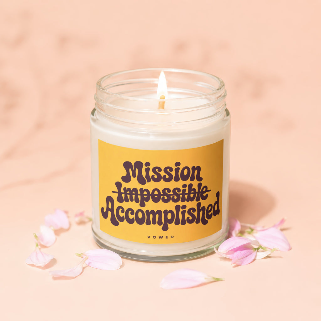 Candle with lit wick, "Mission Impossible Accomplished" text on yellow label, surrounded by petals.