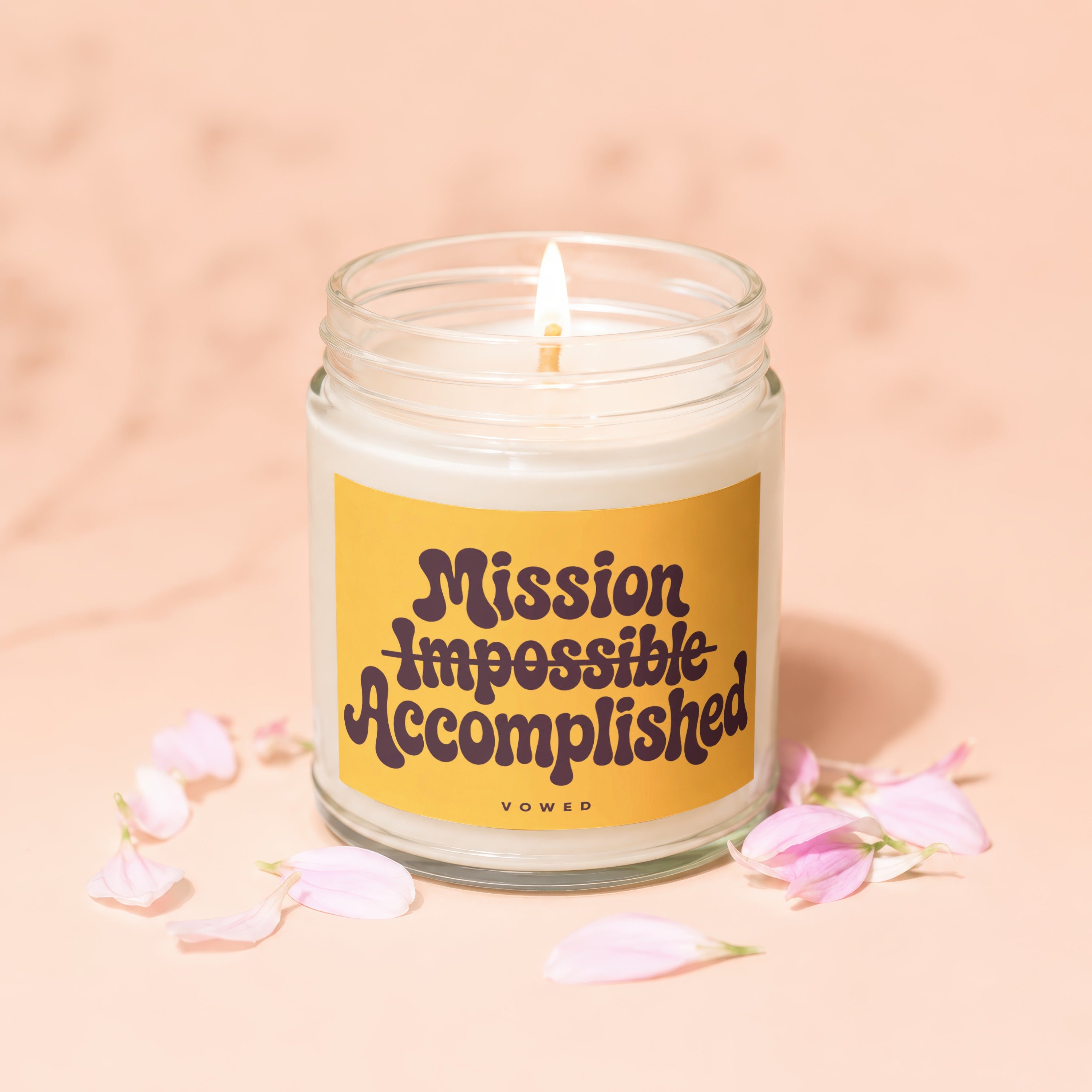 Candle with lit wick, "Mission Impossible Accomplished" text on yellow label, surrounded by petals.
