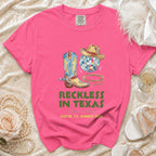 Bright pink t-shirt with cowboy boots, disco ball, hat, and "Reckless in Texas" text.