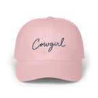 A pink baseball cap with the word cowgirl embroidered in navy blue cursive script on the front.
