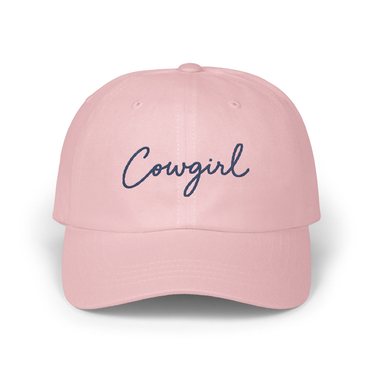 A pink baseball cap with the word cowgirl embroidered in navy blue cursive script on the front.