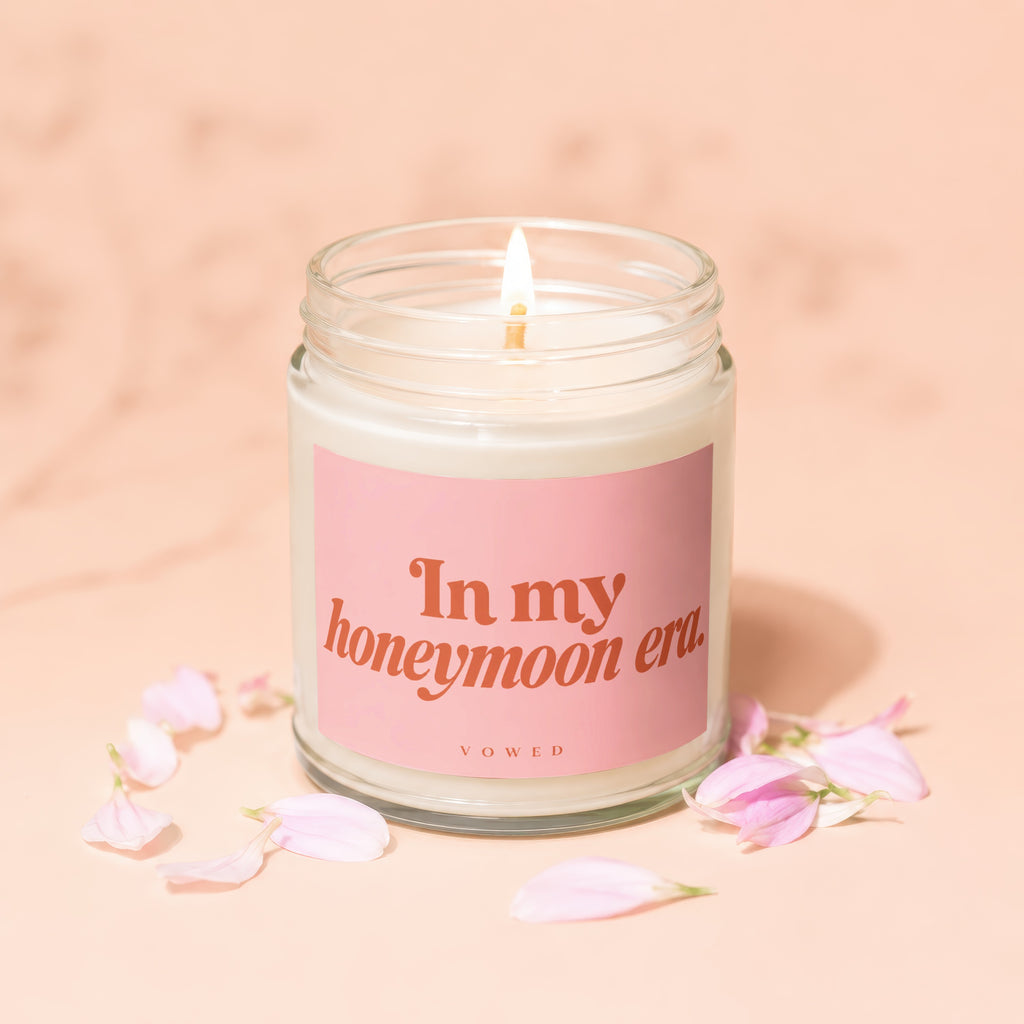 Lit candle with pink label reading "In my honeymoon era VOWED" among pink petals.