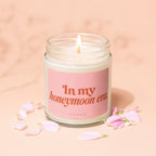 Lit candle with pink label reading "In my honeymoon era VOWED" among pink petals.