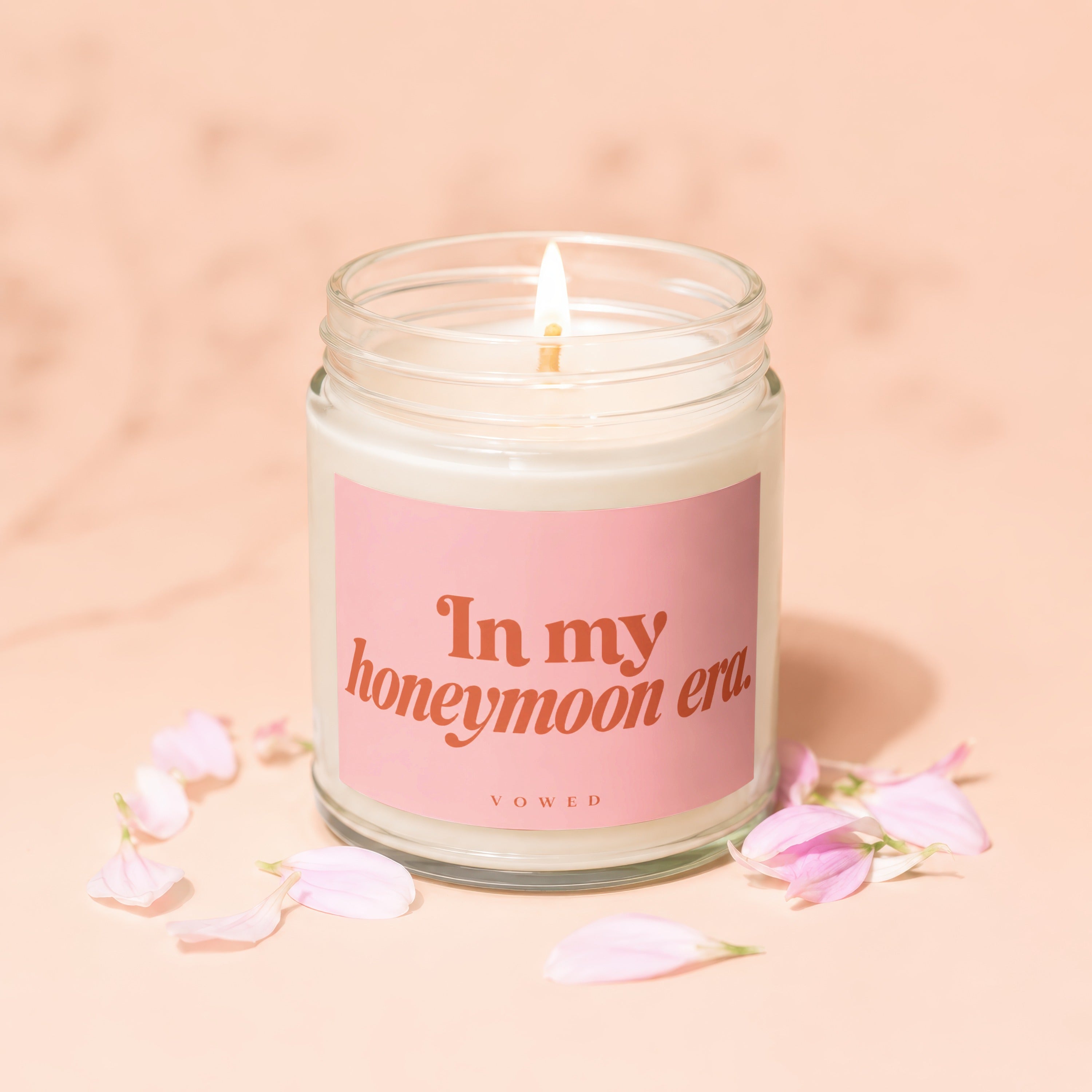 Lit candle with pink label reading "In my honeymoon era VOWED" among pink petals.