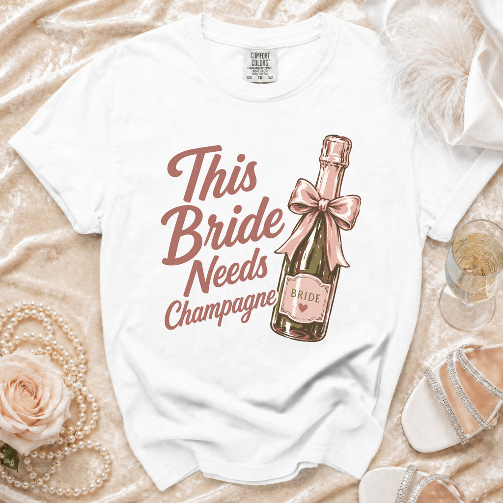 White t-shirt with "This Bride Needs Champagne" text and champagne bottle graphic.