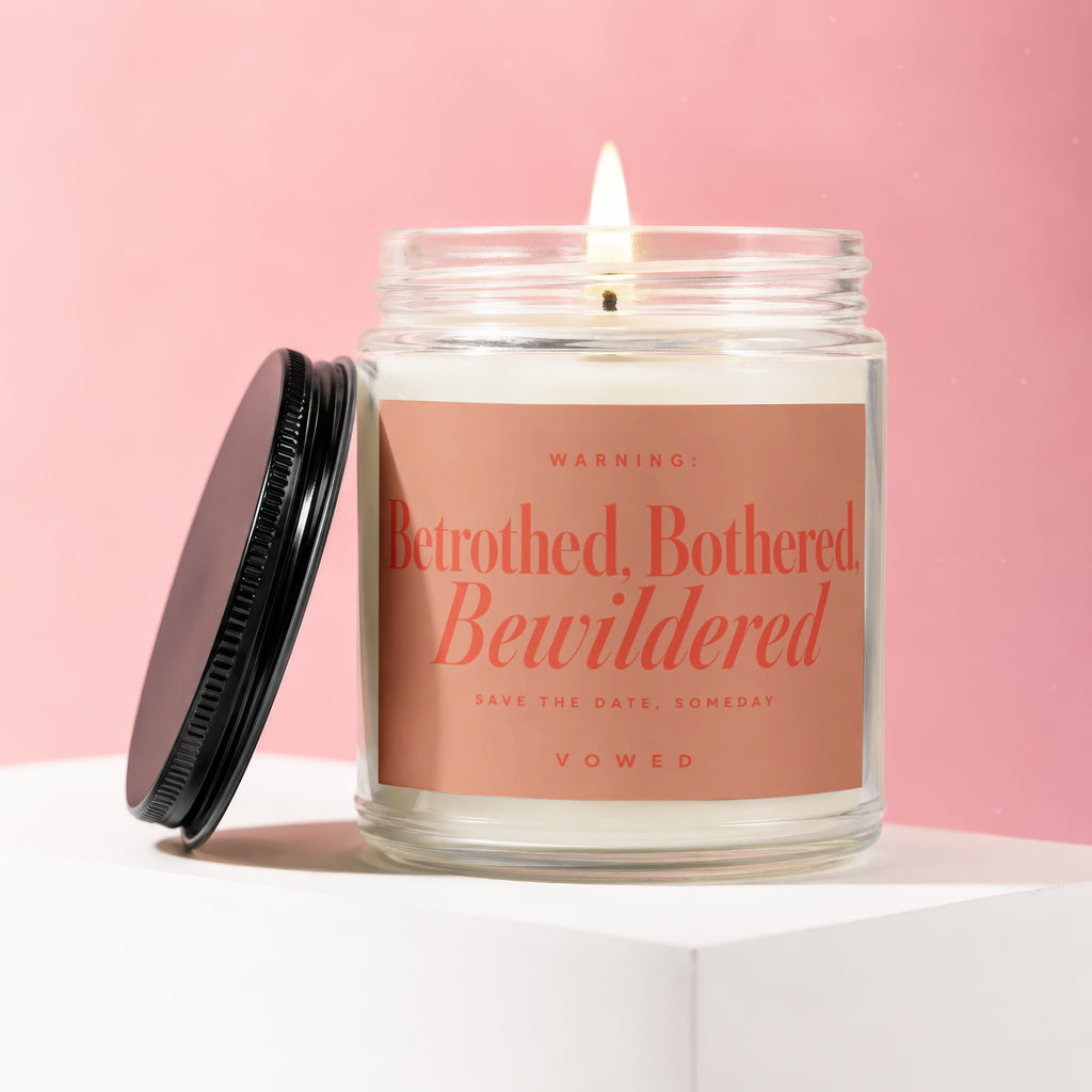 Glass jar candle with lit wick and black lid, reading "Betrothed, Bothered, Bewildered Vowed".