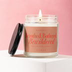 Glass jar candle with lit wick and black lid, reading "Betrothed, Bothered, Bewildered Vowed".