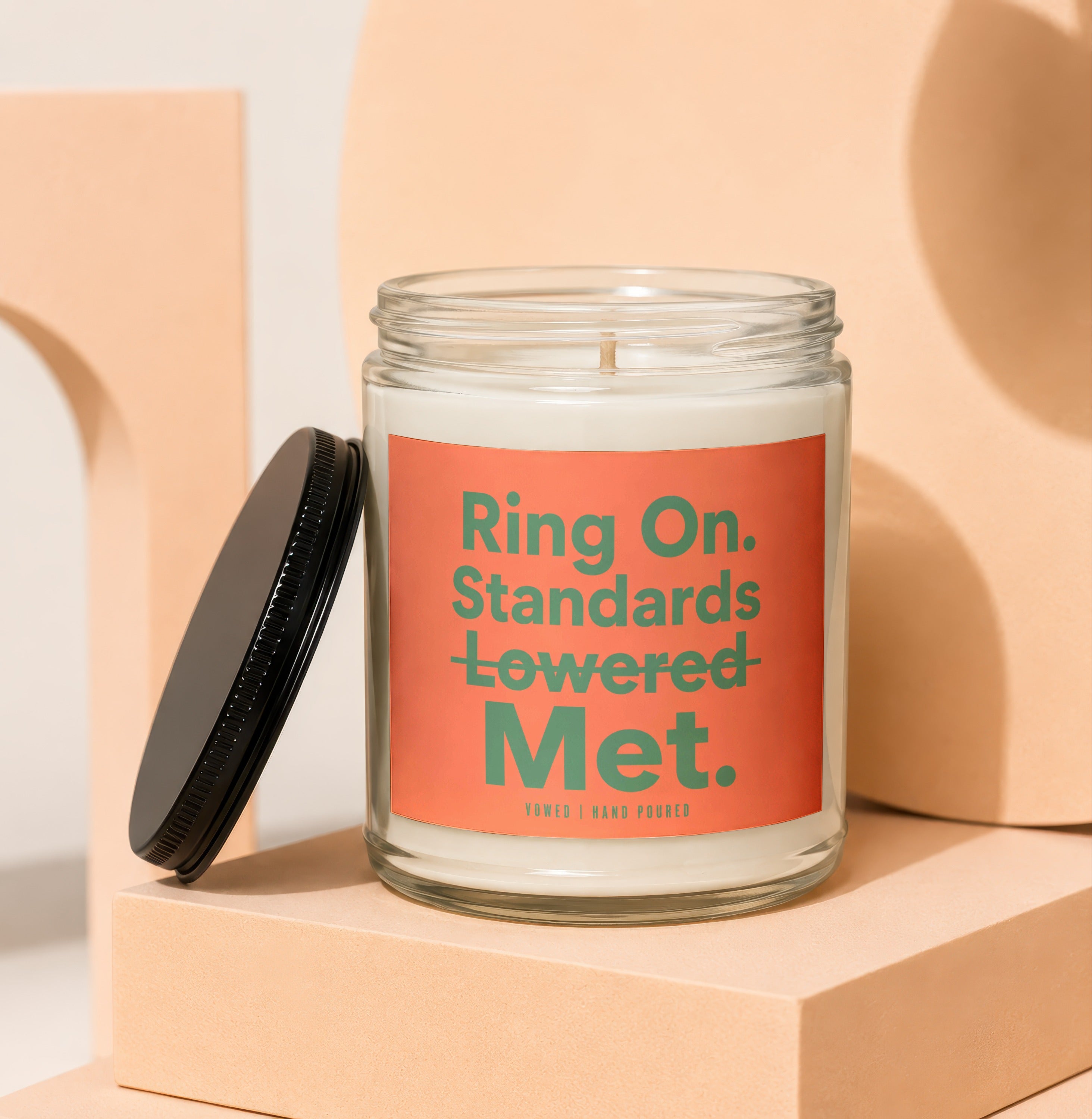 Glass jar candle with orange label reading "Ring On. Standards Lowered Met."