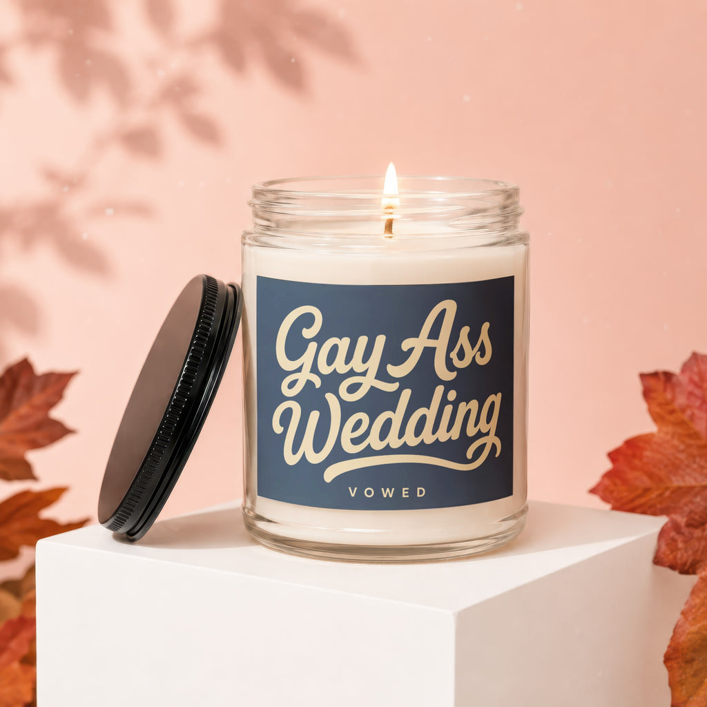 Glass candle with "Gay Ass Wedding" text, lid off, lit wick, autumn leaves.