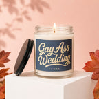 Glass candle with "Gay Ass Wedding" text, lid off, lit wick, autumn leaves.