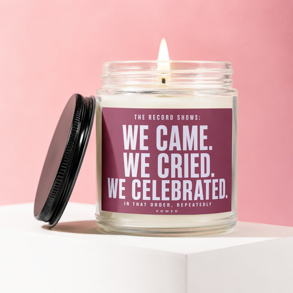 Lit candle in a glass jar with a maroon label reading "We Came. We Cried. We Celebrated."