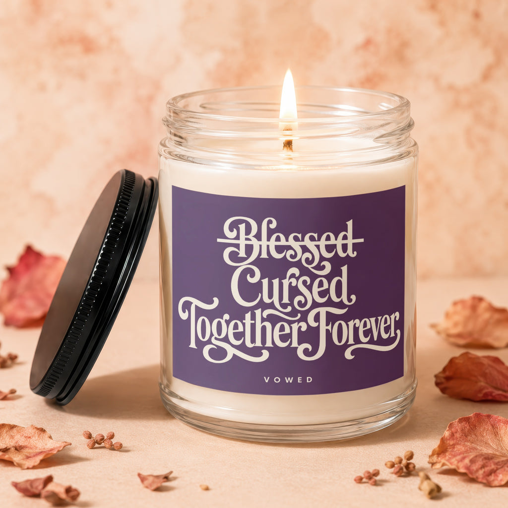 Glass candle with flickering flame on purple label reading "Blessed Cursed Together Forever Vowed."
