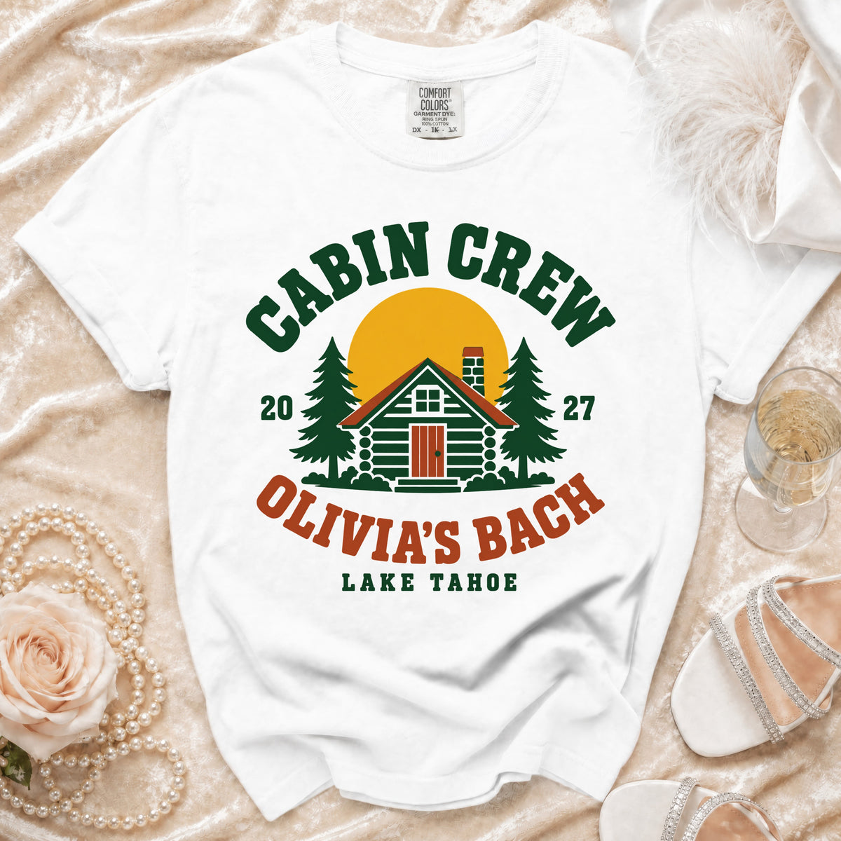 White t-shirt with a green, red, and yellow graphic featuring a cabin, trees, and the text Cabin Crew Olivia's Bach Lake Tahoe.