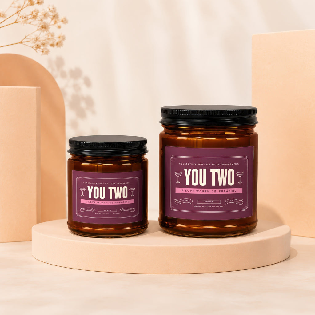 Two amber glass candles with dark lids, featuring "YOU TWO" labels for engagement celebration.