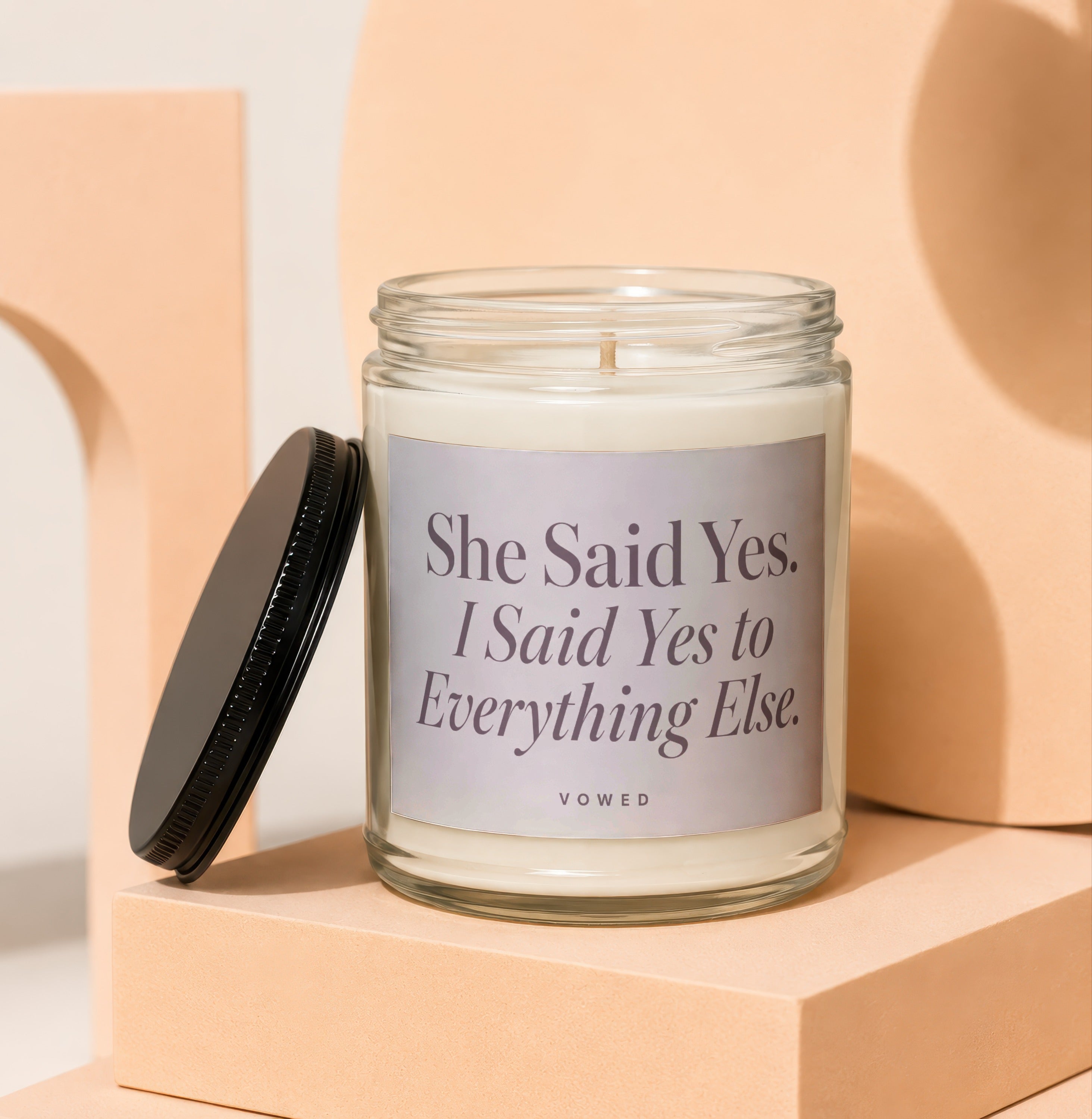 Glass candle with black lid off, labeled "She Said Yes. I Said Yes to Everything Else. VOWED."