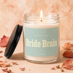 Candle labeled "Smells Like Bride Brain" with lid nearby, surrounded by dried leaves.