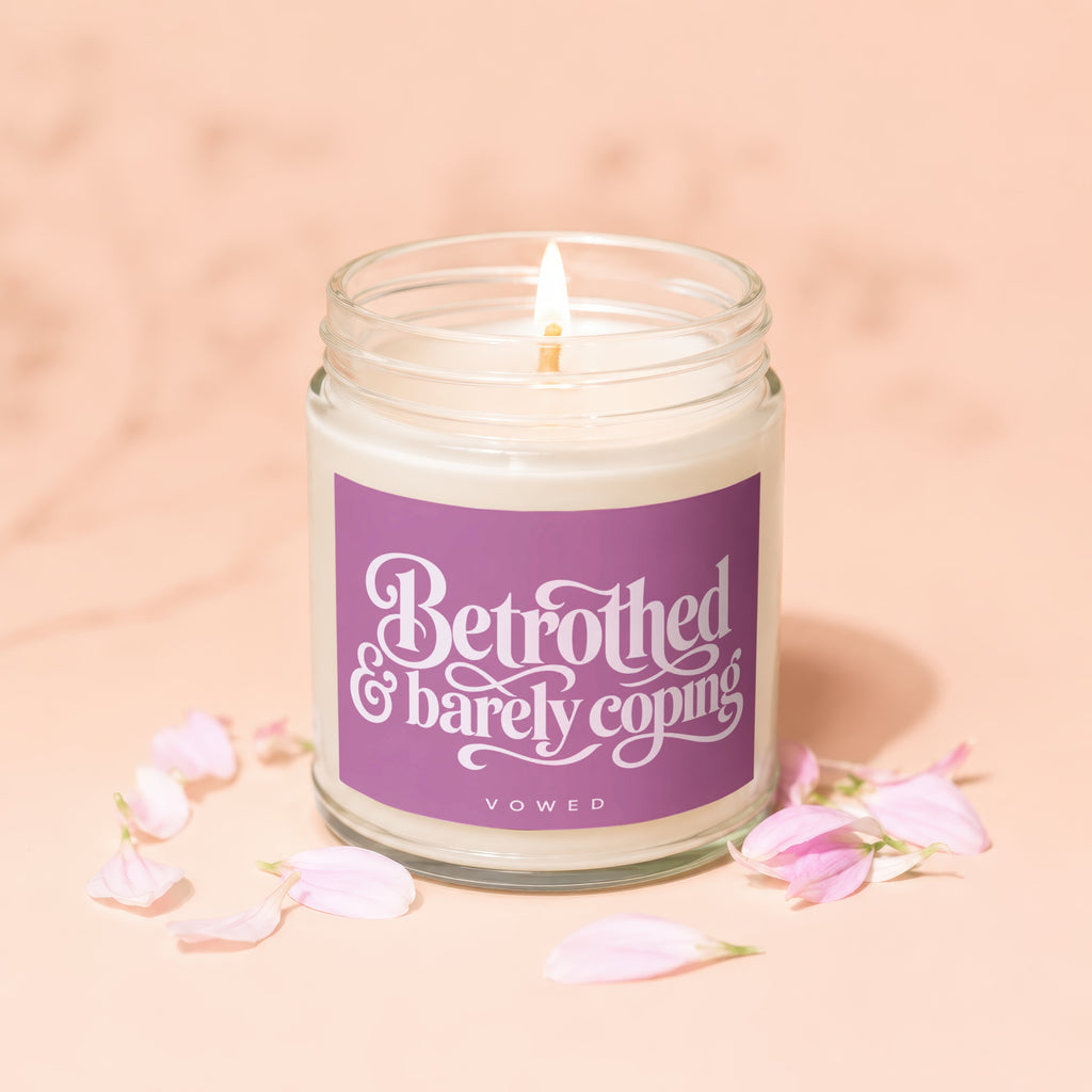 Candle with purple label reading "Betrothed & barely coping" surrounded by pink petals.