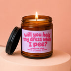 Amber jar candle with a lit wick and a black lid, labeled "will you hold my dress while I pee?".