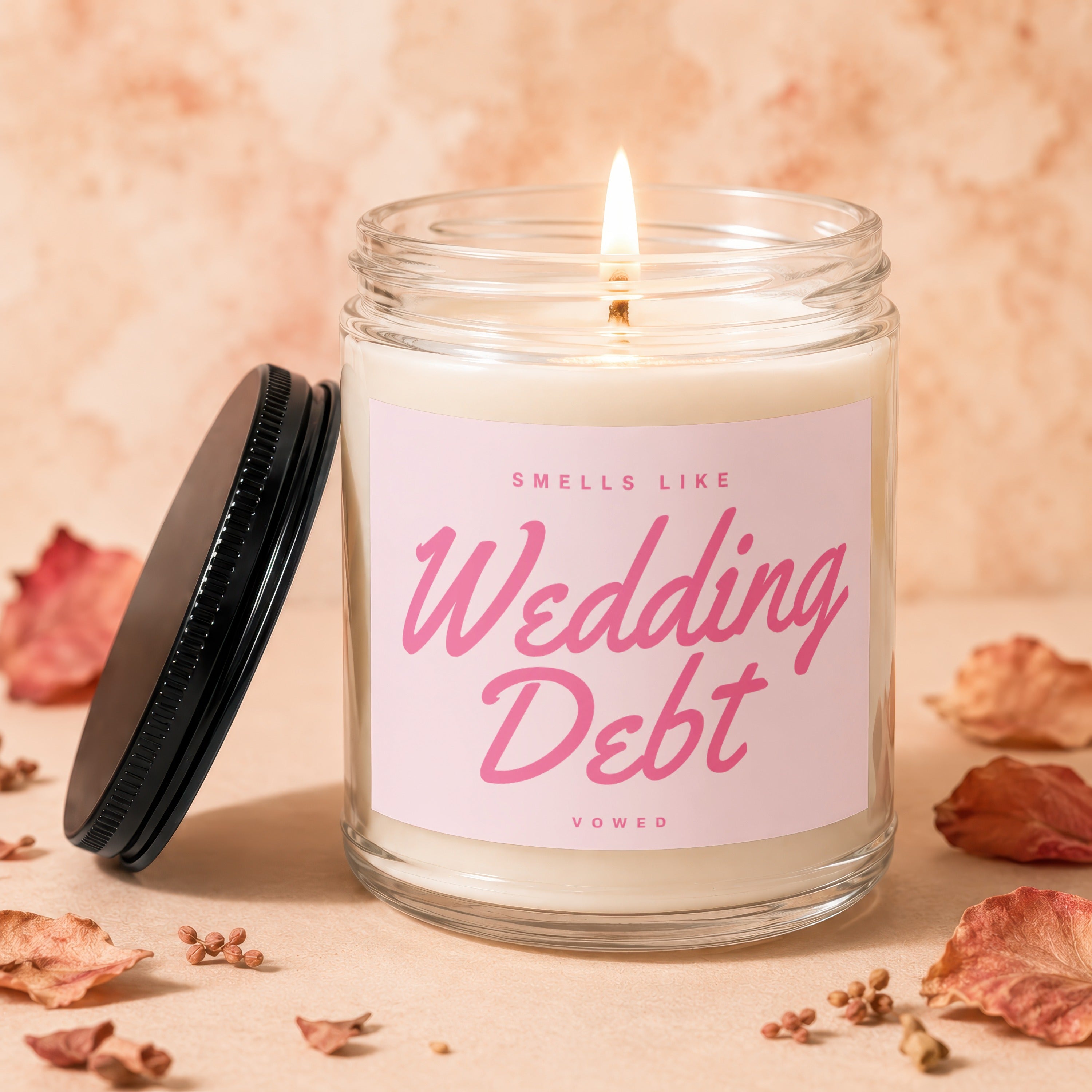 Glass candle with lit wick and pink label reading "Smells Like Wedding Debt Vowed".