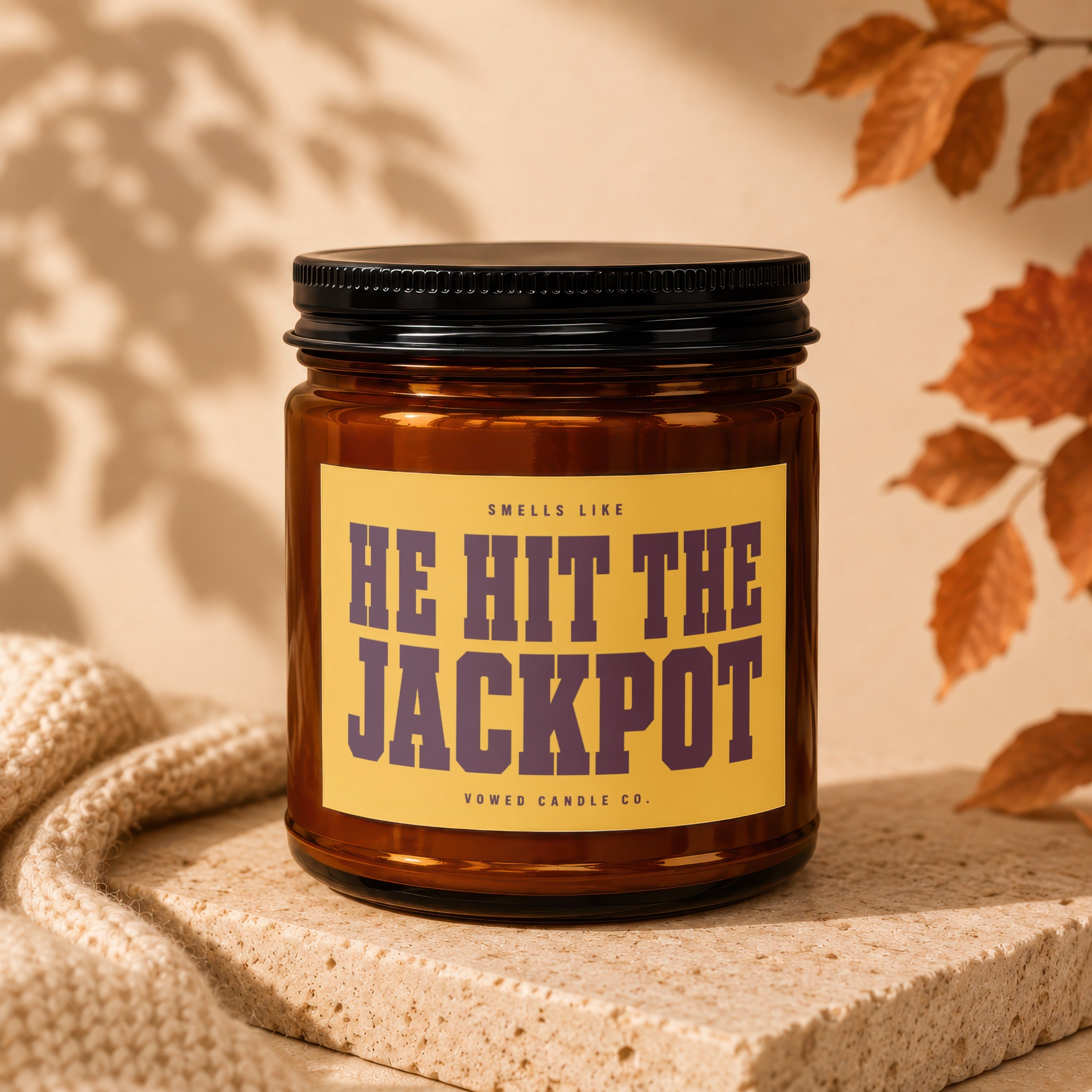 Amber candle with black lid displays a yellow label reading "HE HIT THE JACKPOT" by Vowed Candle Co.