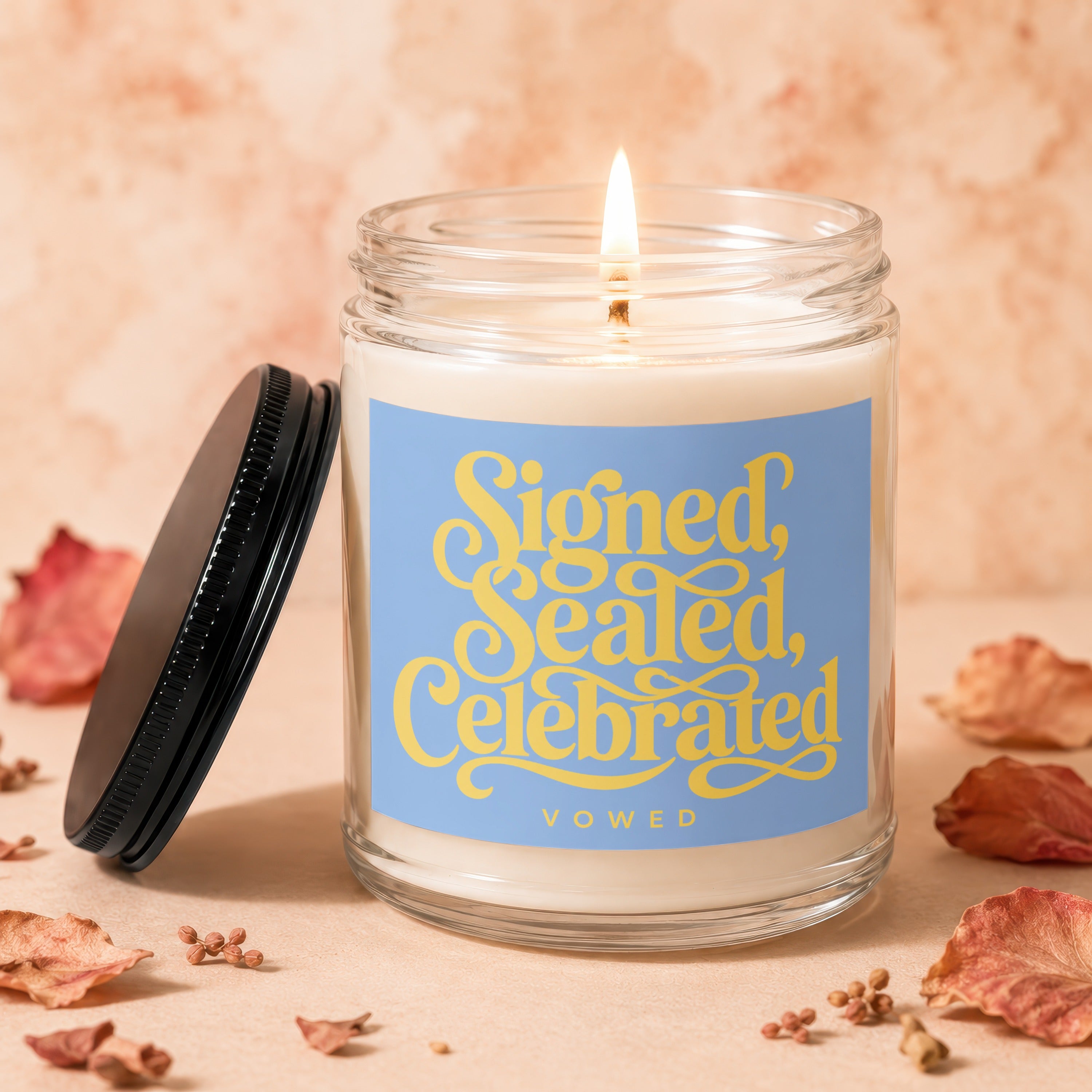 Glass jar candle with lit wick, blue label says "Signed, Sealed, Celebrated VOWED".