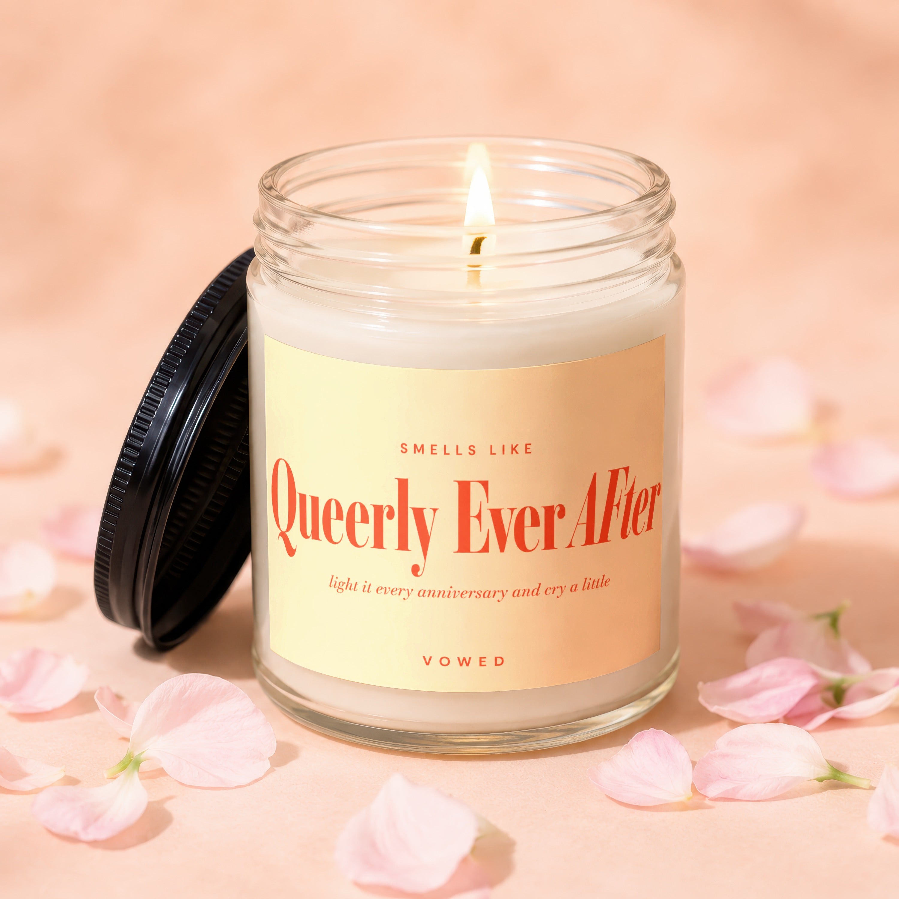 Glass jar candle with lit wick, "Queerly Ever After" text, surrounded by pink petals.