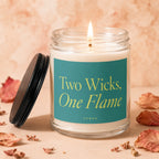 Glass candle with lit wick, 'Two Wicks, One Flame' text, and black lid.