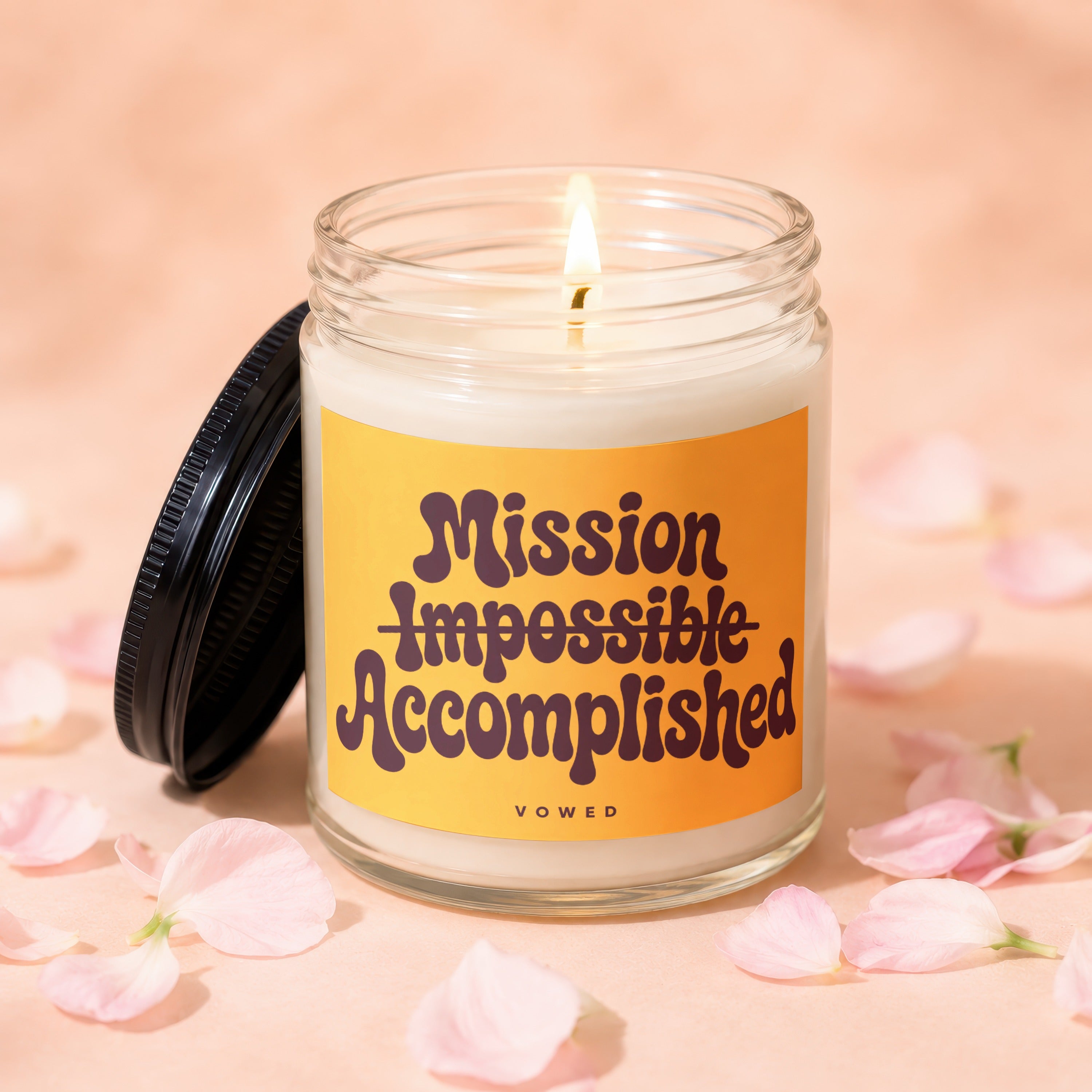 Glass candle with lit wick and yellow label reading "Mission Impossible Accomplished".