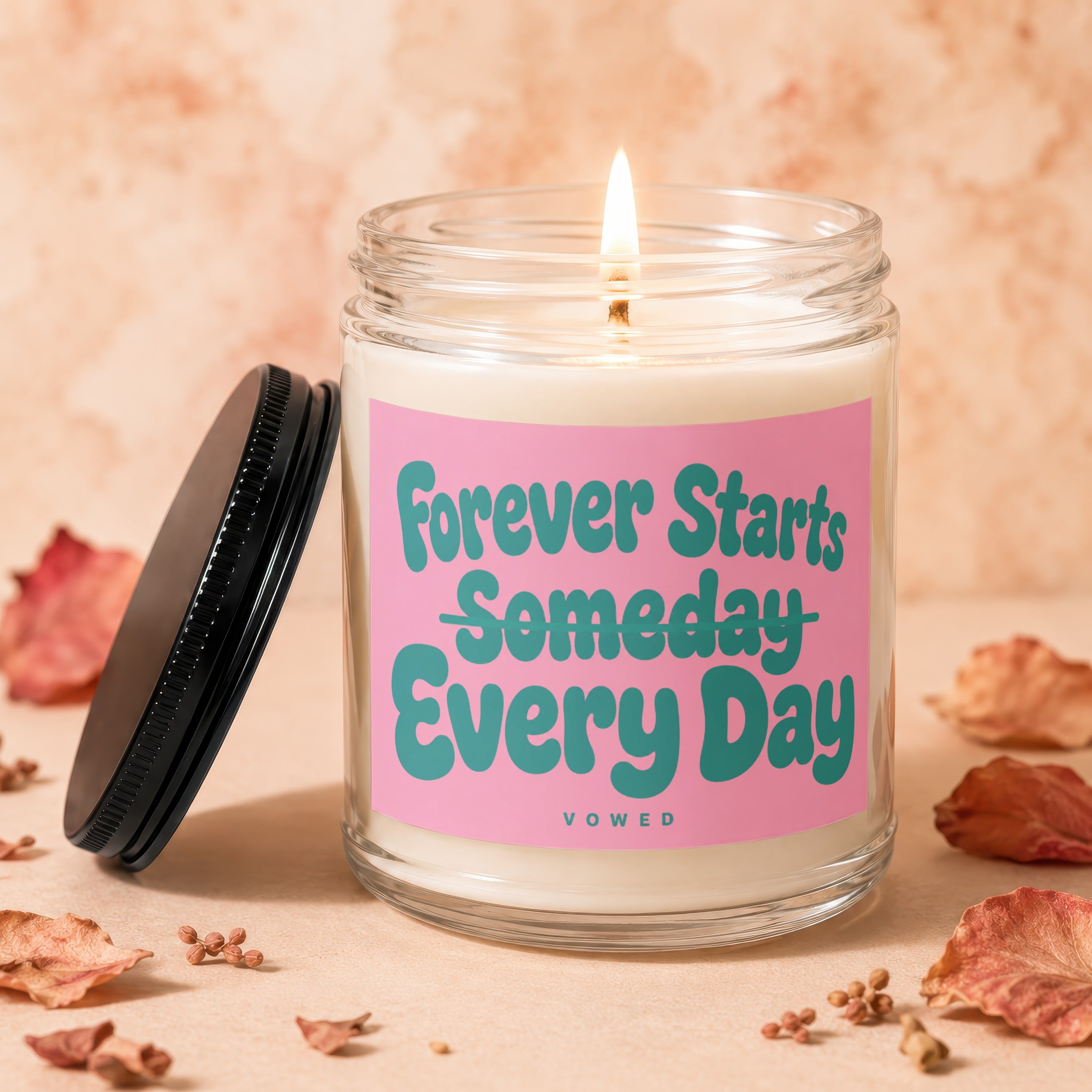 Glass candle with lit wick and pink label reading "Forever Starts Someday Every Day"