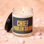 Glass jar candle with burning wick, black lid, and label reading "Chief Problem Solver".