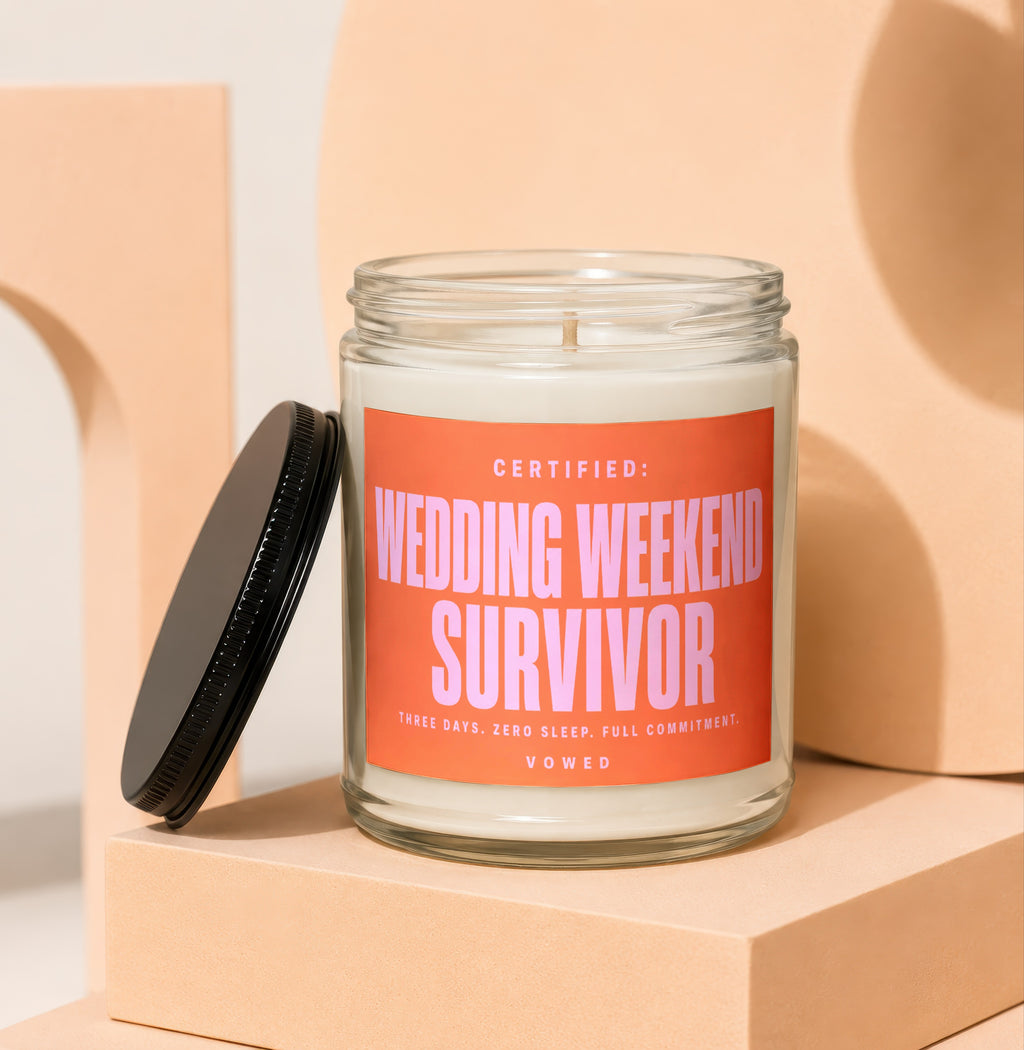 Glass candle with a pink label reading "Certified: Wedding Weekend Survivor. Three Days. Zero Sleep. Full Commitment. Vowed."