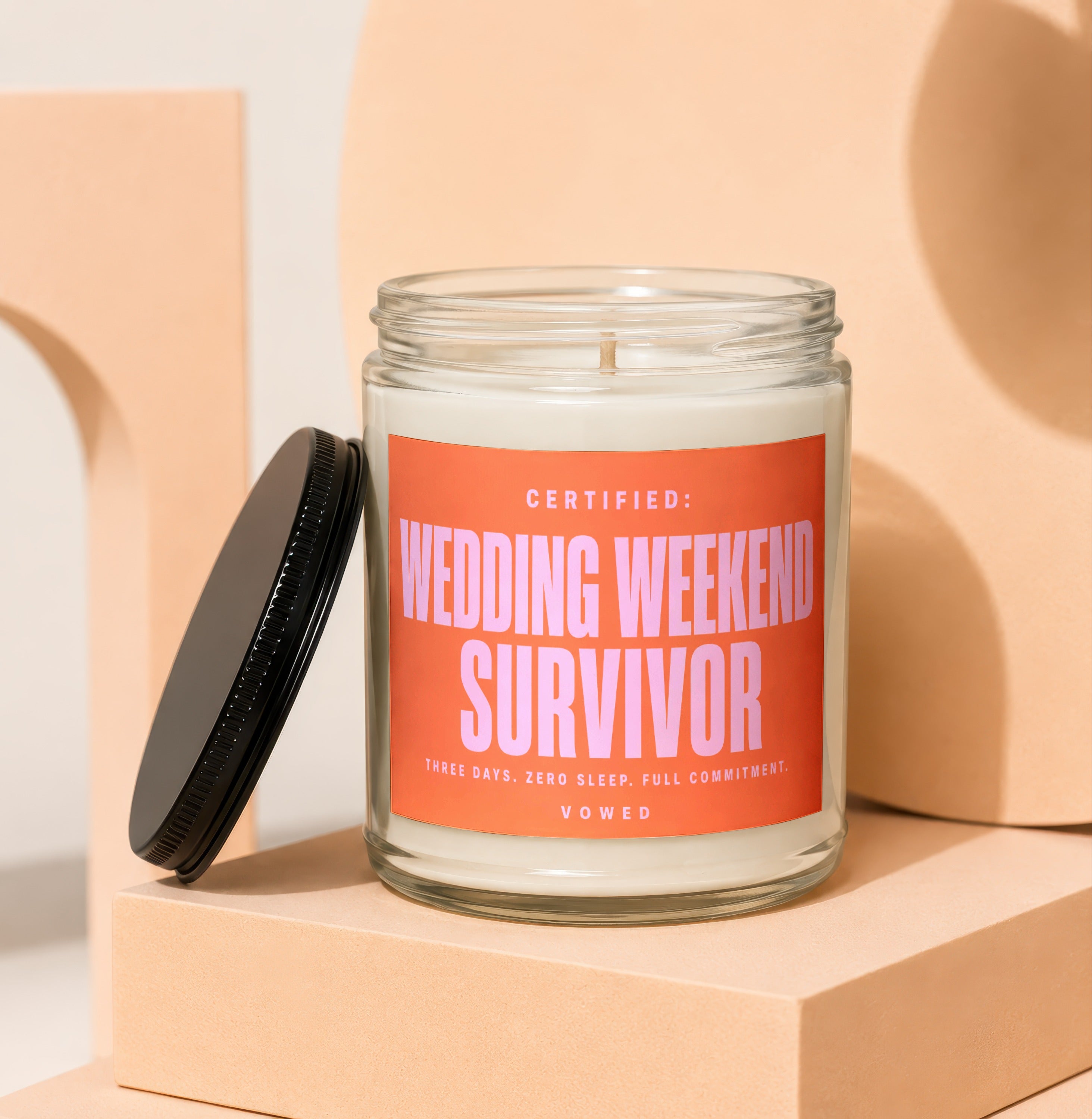 Glass candle with a pink label reading "Certified: Wedding Weekend Survivor. Three Days. Zero Sleep. Full Commitment. Vowed."