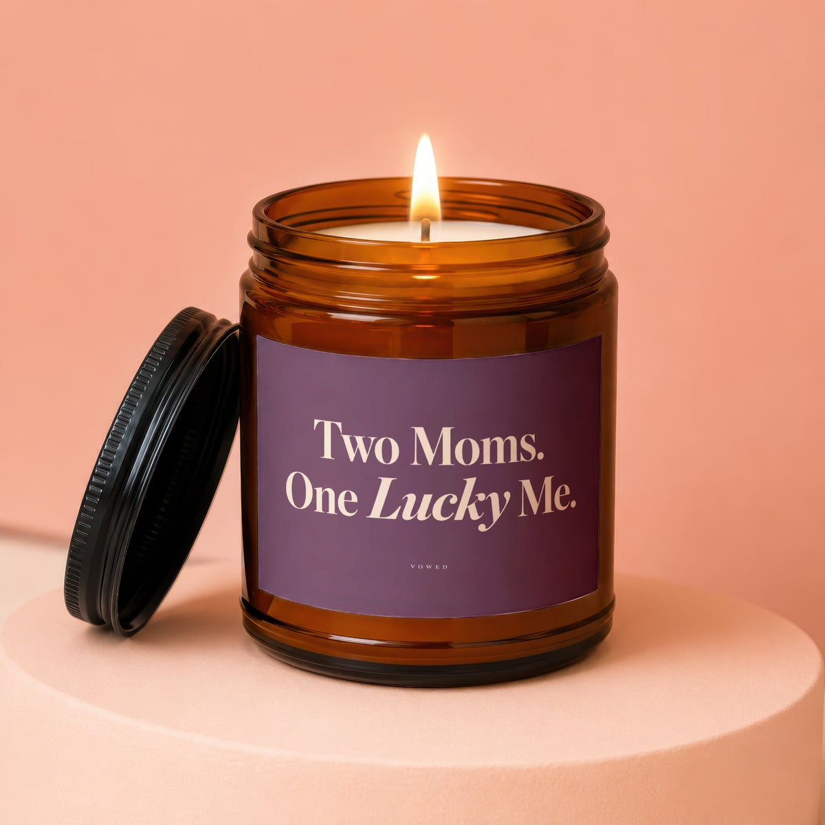 Amber jar candle with lit wick and lid off, reads "Two Moms. One Lucky Me."