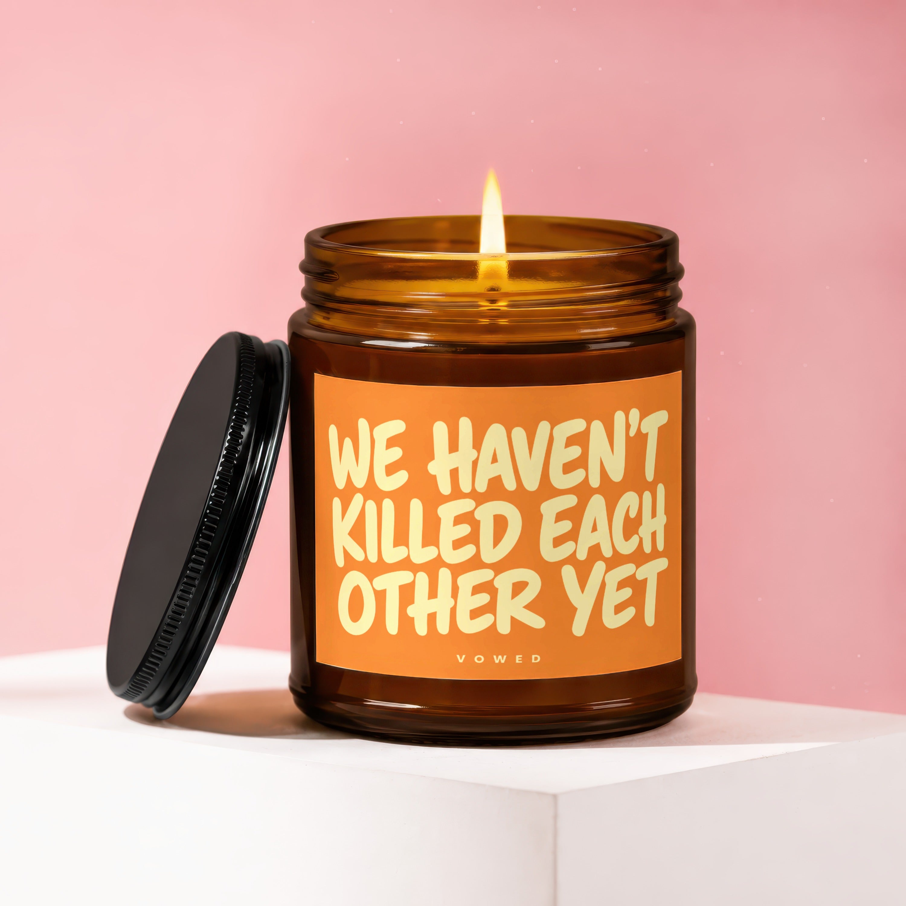 Lit amber glass candle with orange label reading "WE HAVEN'T KILLED EACH OTHER YET".