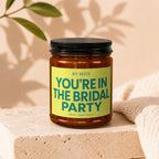 Glass candle with black lid, yellow label reading "HEY BESTIE YOU'RE IN THE BRIDAL PARTY".