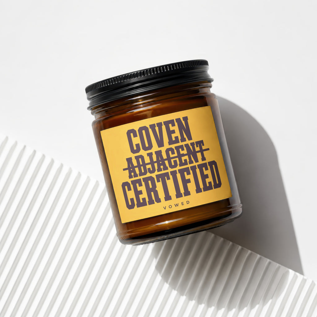 Amber glass candle with black lid and yellow label reading "COVEN ADJACENT CERTIFIED".