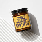 Amber glass candle with black lid and yellow label reading "COVEN ADJACENT CERTIFIED".