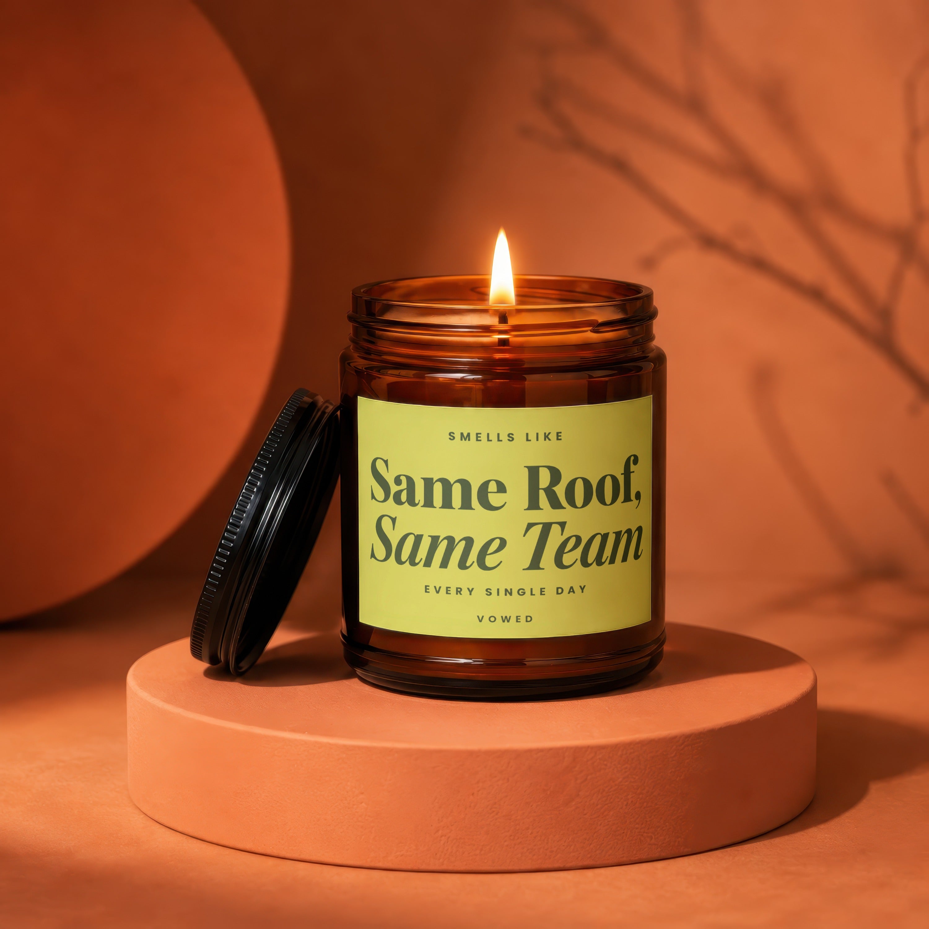 Amber candle with lit wick reads "Smells Like Same Roof, Same Team."