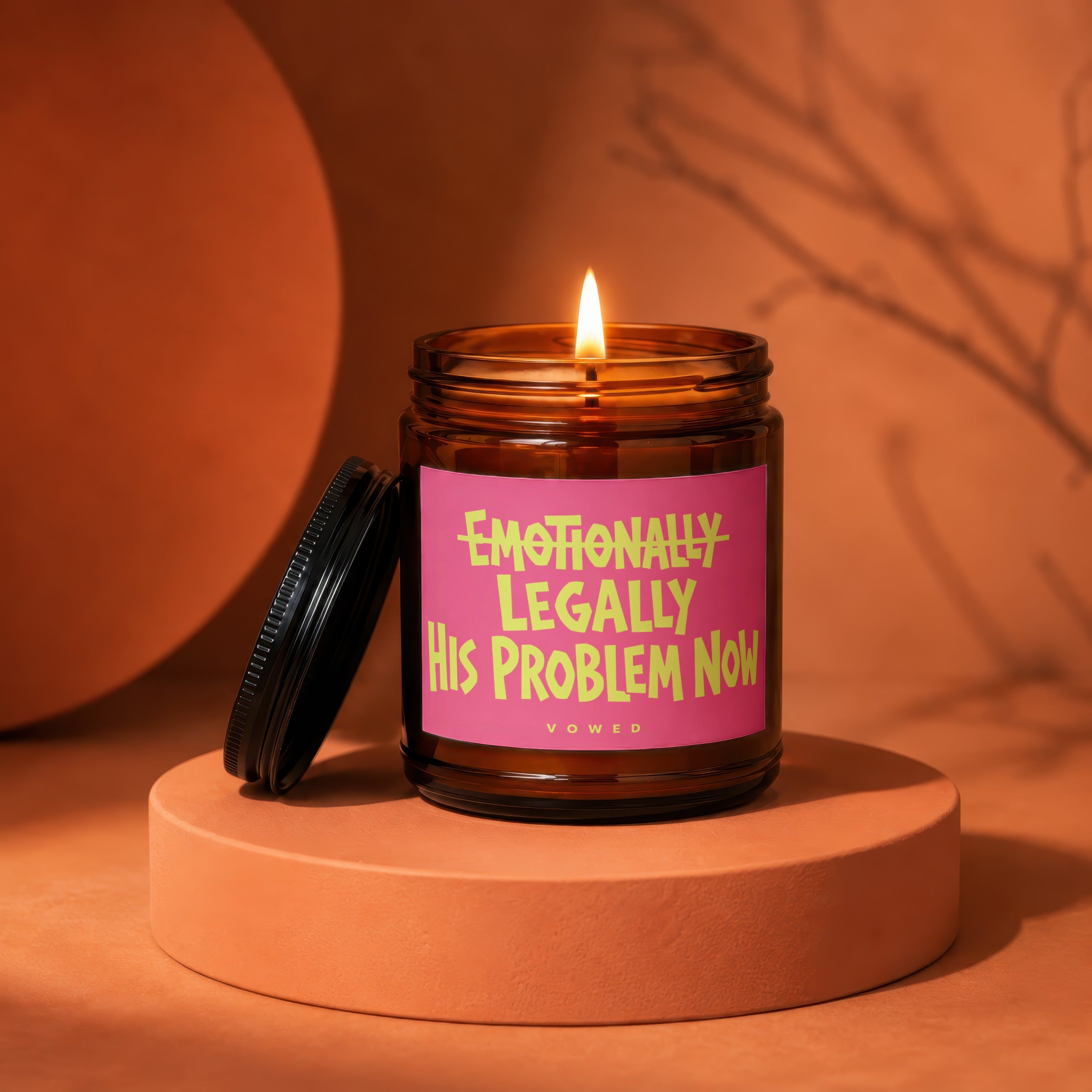 Amber glass candle with lit flame and pink label reading "Emotionally Legally His Problem Now".