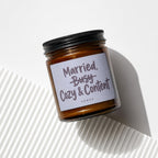 Amber jar candle with "Married, Busy, Cozy & Content" text, on white surface.
