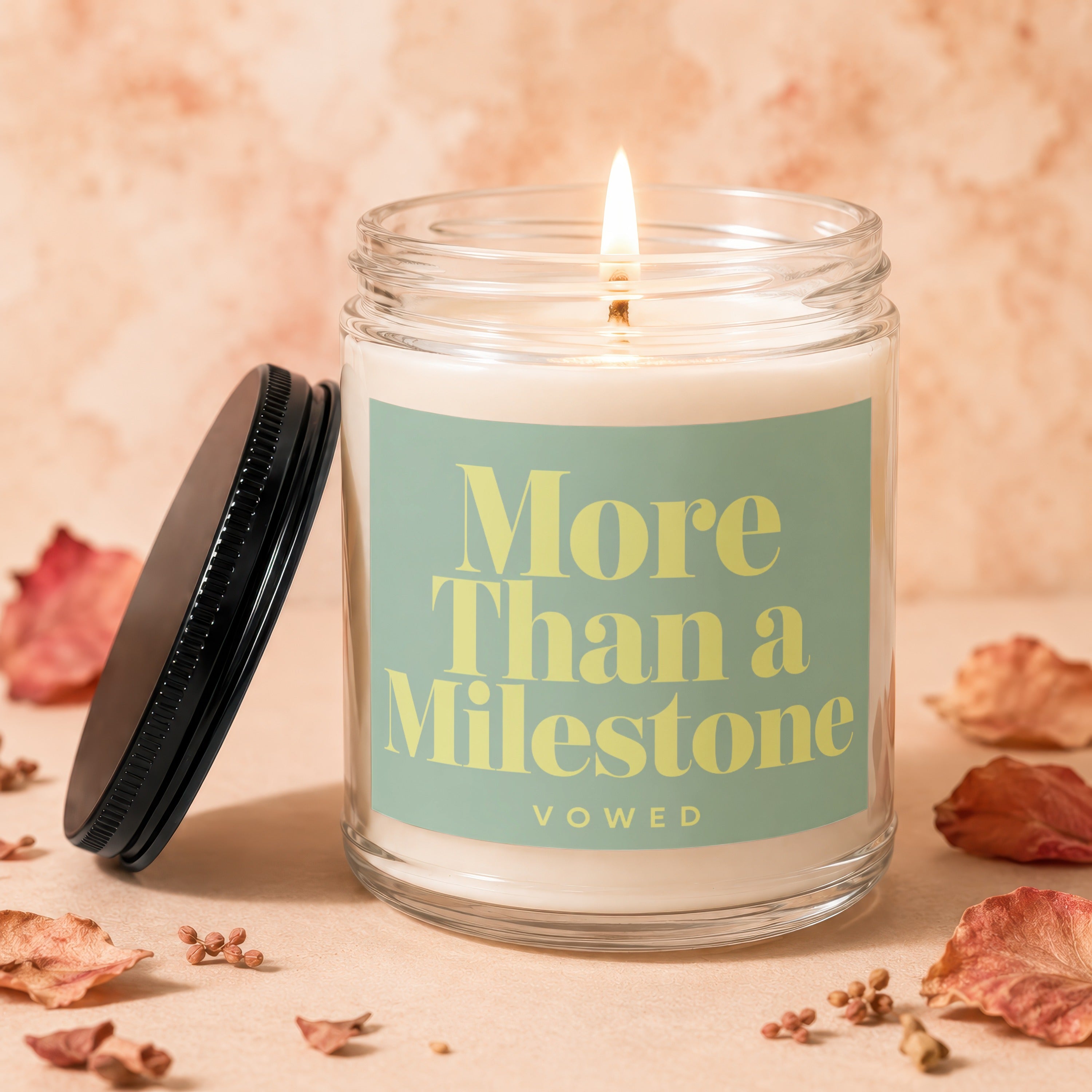 Lit candle in glass jar with "More Than a Milestone" text, lid off.