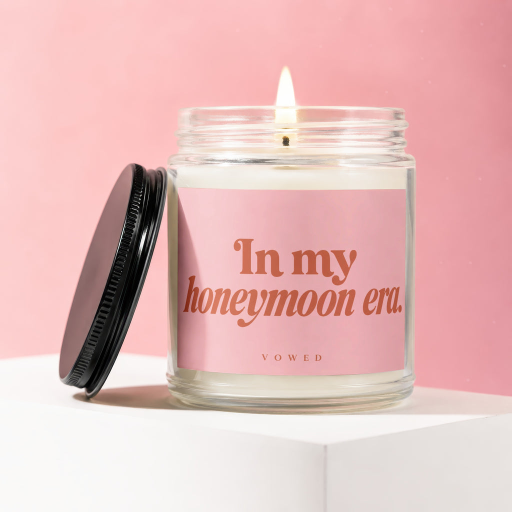 Glass candle with lit wick on pink background, says "In my honeymoon era".