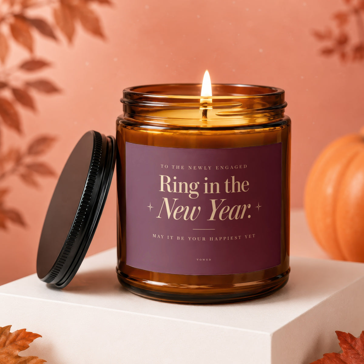 Candle with "Ring in the New Year" text, lit flame, lid removed, and autumn decor.