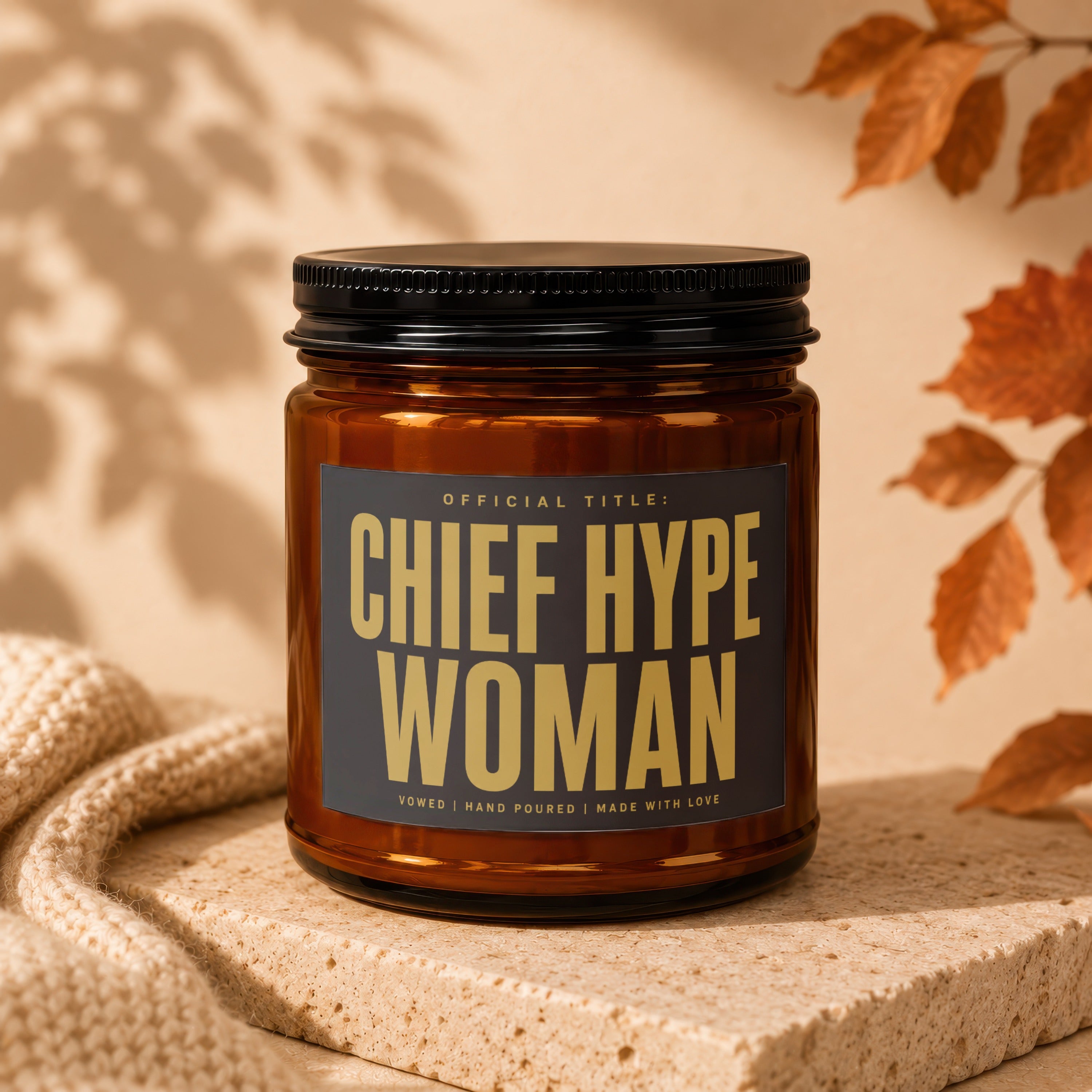 Amber candle jar with black lid, labeled "Chief Hype Woman," rests on speckled stone.