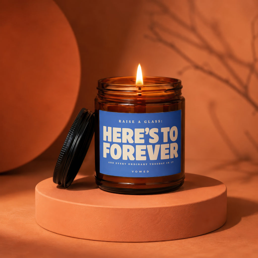 Amber jar candle with lit flame, blue label reads "Here's To Forever" and "Vowed".