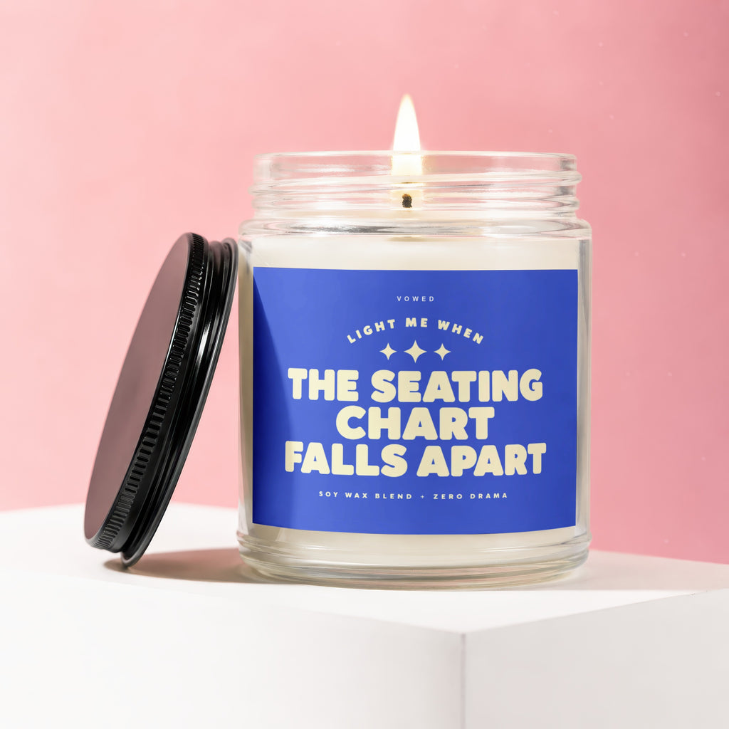 Glass candle with lit flame, featuring label text "The Seating Chart Falls Apart".