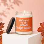 Candle with orange label reading "Welcome to My Budget Wedding" on a white cube.